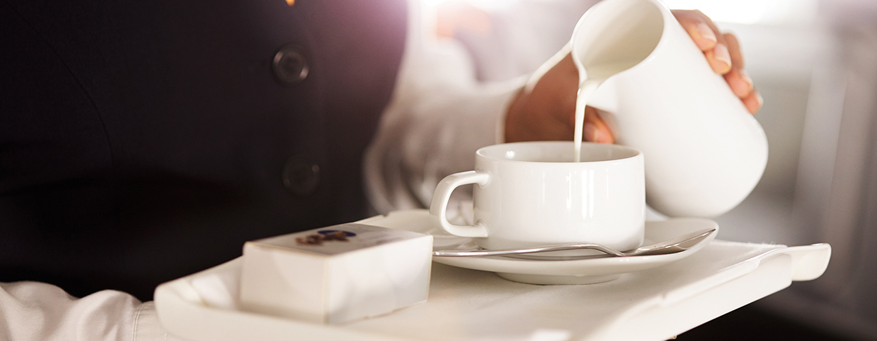 Nahaufnahme eines Tabletts in den Händen einer Flugbegleiterin, welche Milch in eine Tasse Kaffee gießt